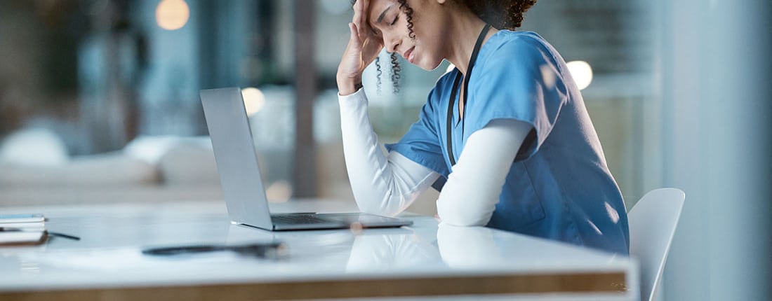 A stressed-out nurse sits in front of a laptop with her head in her hands