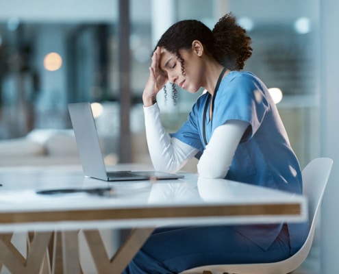 A stressed-out nurse sits in front of a laptop with her head in her hands