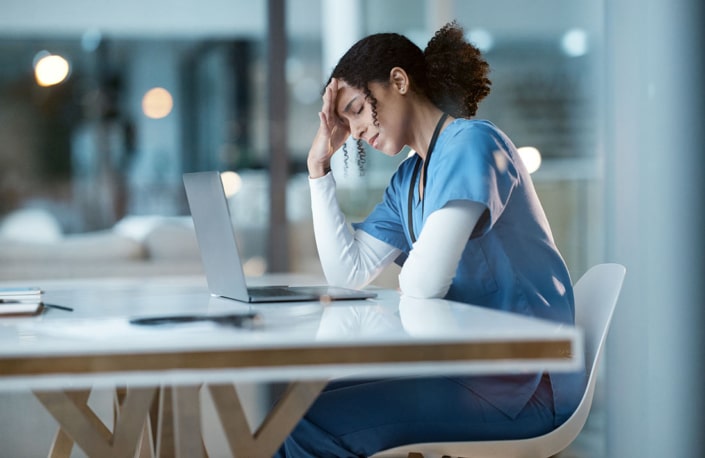 A stressed-out nurse sits in front of a laptop with her head in her hands
