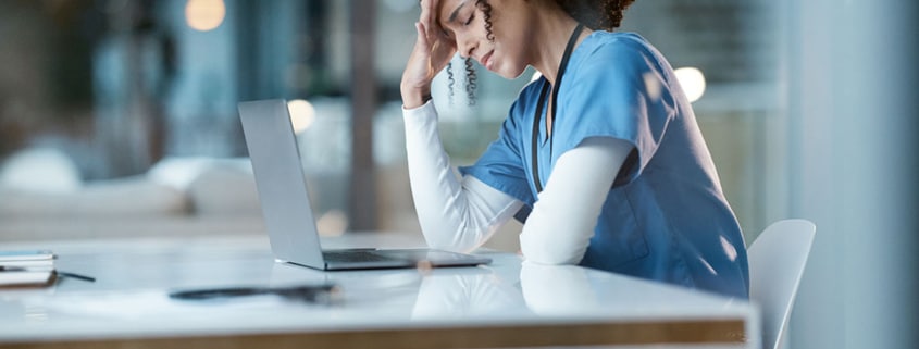 A stressed-out nurse sits in front of a laptop with her head in her hands