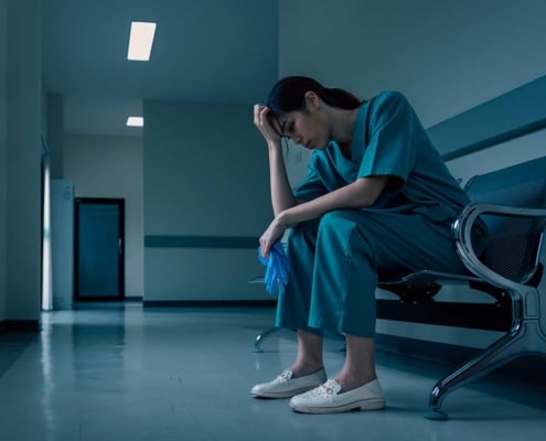 A burned-out nurse sits in a waiting room chair with her head in her hands